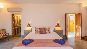 Habitación de hotel con cama, dos lámparas de noche, baño visible a través de una puerta abierta y un banco de madera.