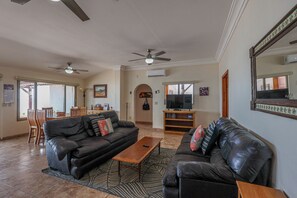 A spacious living room with a black leather sofa, a wooden coffee table, and a television. There is a dining area with chairs and a table, and a ceiling fan. - Casa Evita – Beachfront, Casita, Rocky Point (Puerto Penasco)