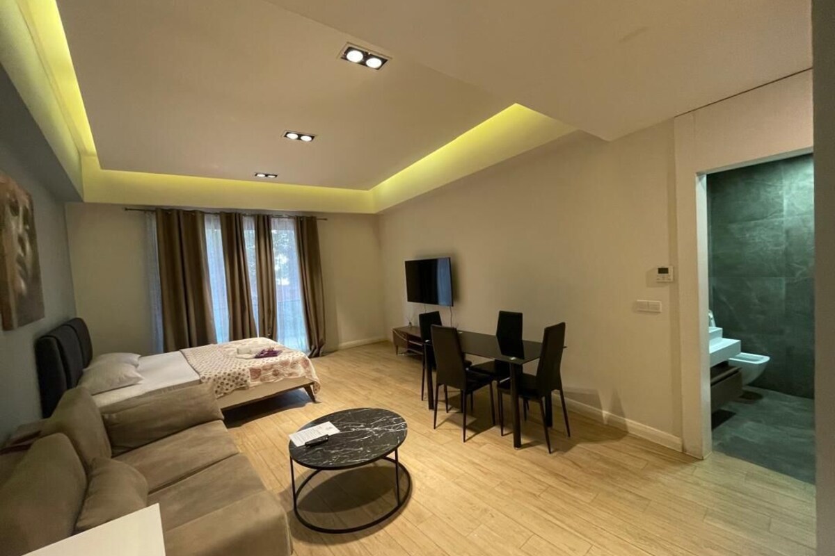 Ein modernes Hotelzimmer mit Bett, Sofa, Esstisch, Stühlen, einem Flachbildfernseher und einem durchgegangenen Tür sichtbaren Badezimmer.