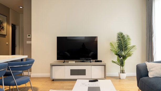 Ein modernes Wohnzimmer mit Flachbildfernseher, einem Essbereich mit blauen Stühlen, einem grauen Sofa und einer Topfpflanze.