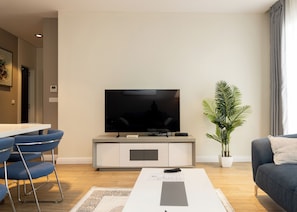 A modern living room with a flat-screen TV, a dining area with blue chairs, a grey sofa, and a potted plant. - The Pearl In City Apartments (Accra)