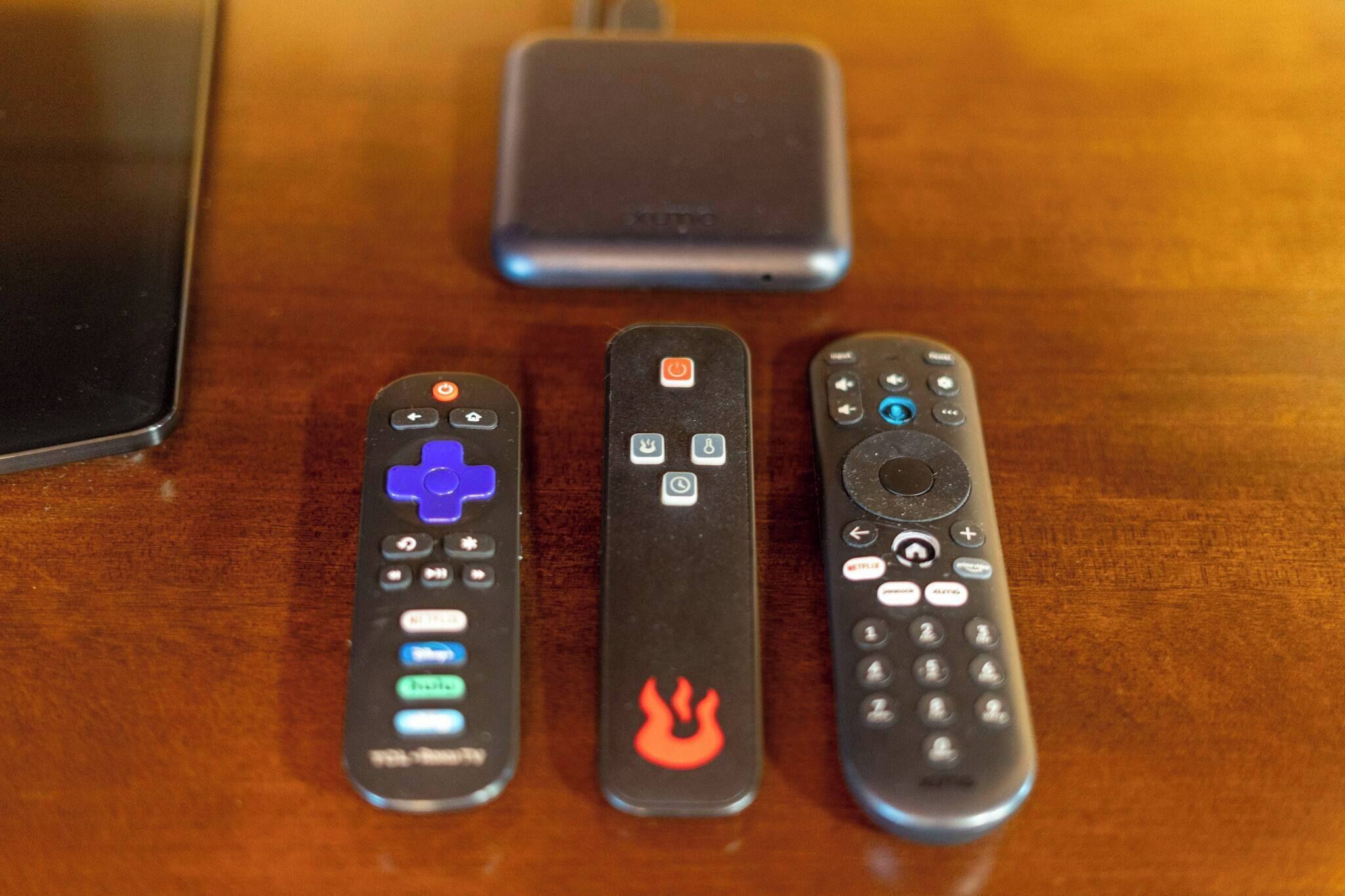 Three remote controls on a wooden surface with a smartphone and a power bank in the background.