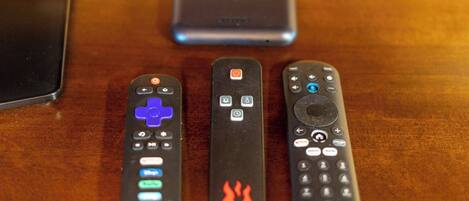 Three remote controls on a wooden surface with a smartphone and a power bank in the background.