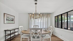 A dining area with a round table, white chairs, a chandelier, and a view through large windows. - Renovated Home Close to Everything! Fenced Yard, Dogs Welcome! (Saint Simons Island)