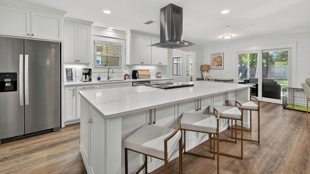 A modern kitchen with white cabinetry, a stainless steel refrigerator, a central island with bar stools, and a large window providing natural light. - Renovated Home Close to Everything! Fenced Yard, Dogs Welcome! (Saint Simons Island)