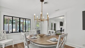 A dining area with a round table, white chairs, a chandelier, and large windows. - Renovated Home Close to Everything! Fenced Yard, Dogs Welcome! (Saint Simons Island)