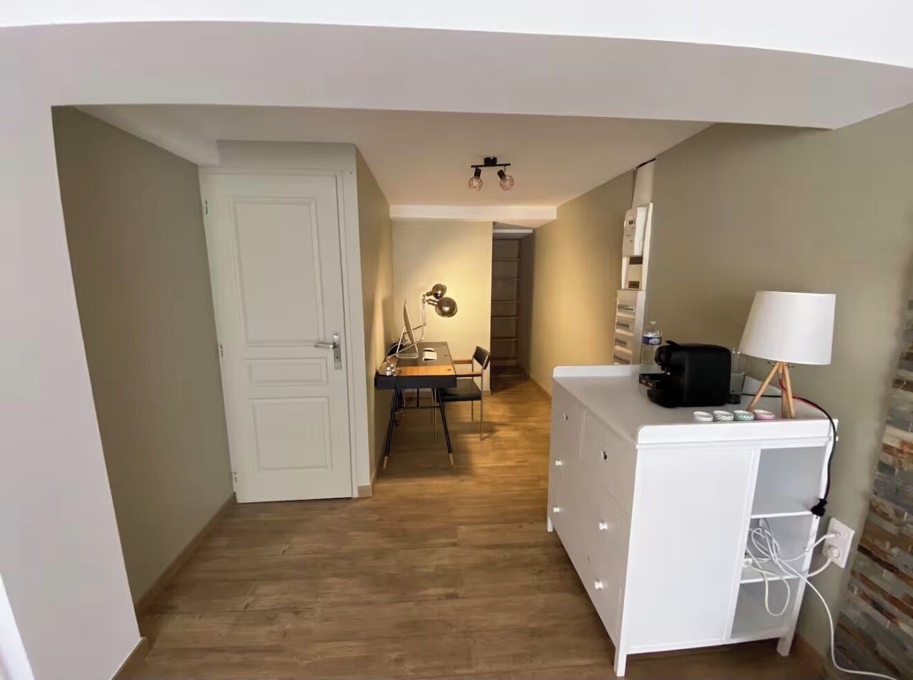 A room with a wooden floor, a white dresser, a desk with a lamp, and a door leading to another room.