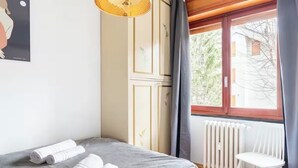 Una camera da letto con un letto, asciugamani arrotolati, un termosifone, una finestra con tende e un lampadario.