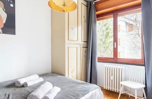 Ein Schlafzimmer mit einem Bett, gerollten Handtüchern, einem Heizkörper, einem Fenster mit Vorhängen und einer Pendelleuchte.