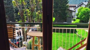 Un balcone con vista su un prato verde, alberi e una montagna. Sono presenti sedie in legno e un tavolo.