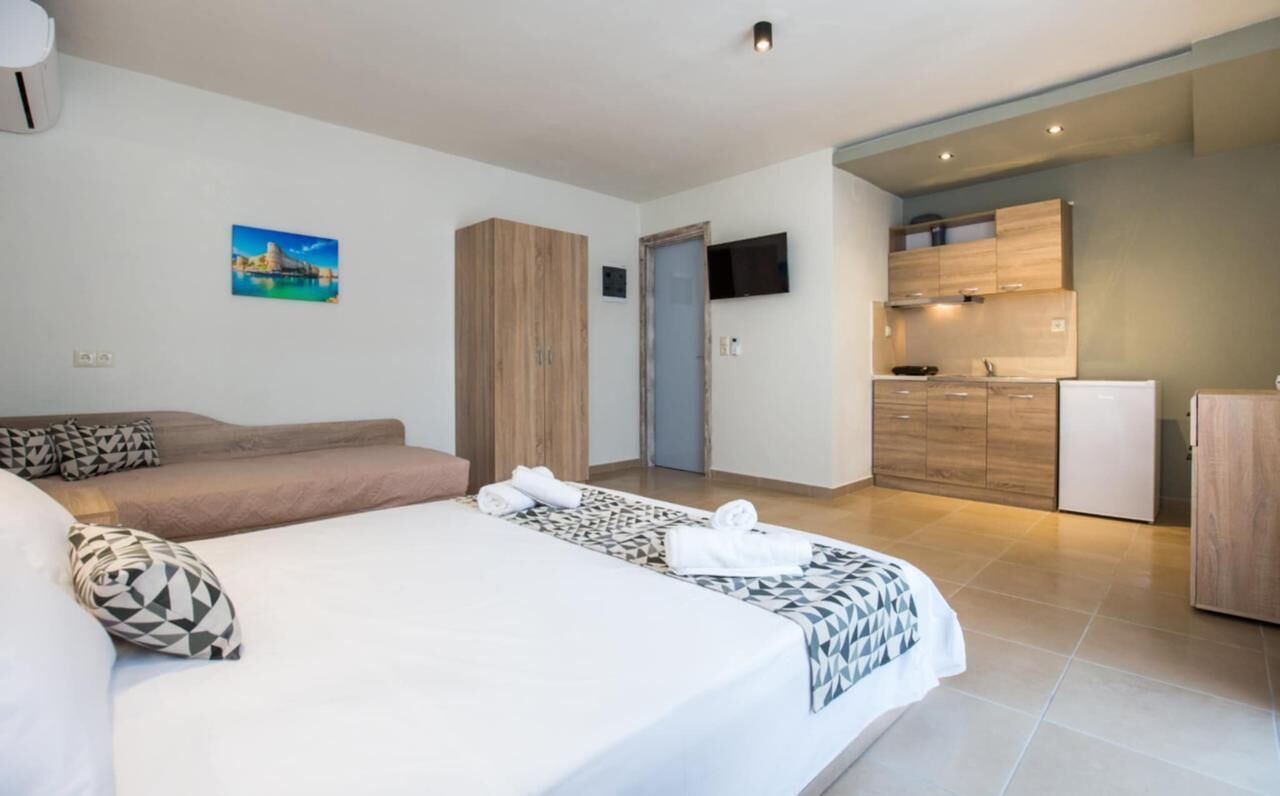 A hotel room with a bed, a sofa, a TV, a kitchen area, and a door.
