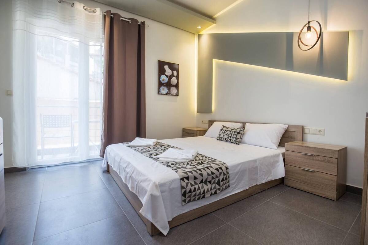 A modern bedroom with a large bed, a bedside table, a nightstand, a wall-mounted light, and a window with curtains.