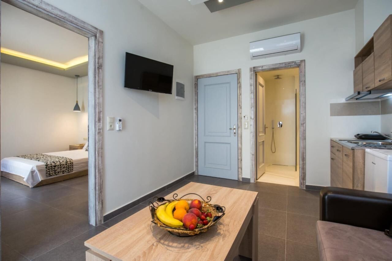 A compact living space with a kitchenette, a dining area with a fruit bowl, a TV mounted on the wall, and a separate bedroom visible through an open door.