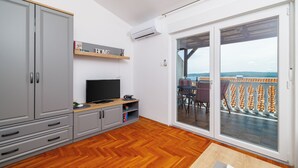 Ein Zimmer mit Fernseher, grauen Schränken, Holzboden und einer Glasschiebetür zum Balkon. An der Wand hängt ein Schild mit der Aufschrift 'HOME'.