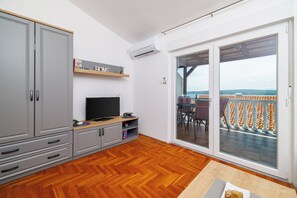 Ein Zimmer mit Fernseher, grauen Schränken, Holzboden und einer Glasschiebetür zum Balkon. An der Wand hängt ein Schild mit der Aufschrift 'HOME'.