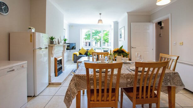 A dining area with a table set for four, a kitchen with a refrigerator and stove, and a living area with a sofa and TV. - Superb 5 Bedroom TownHouse (Galway)