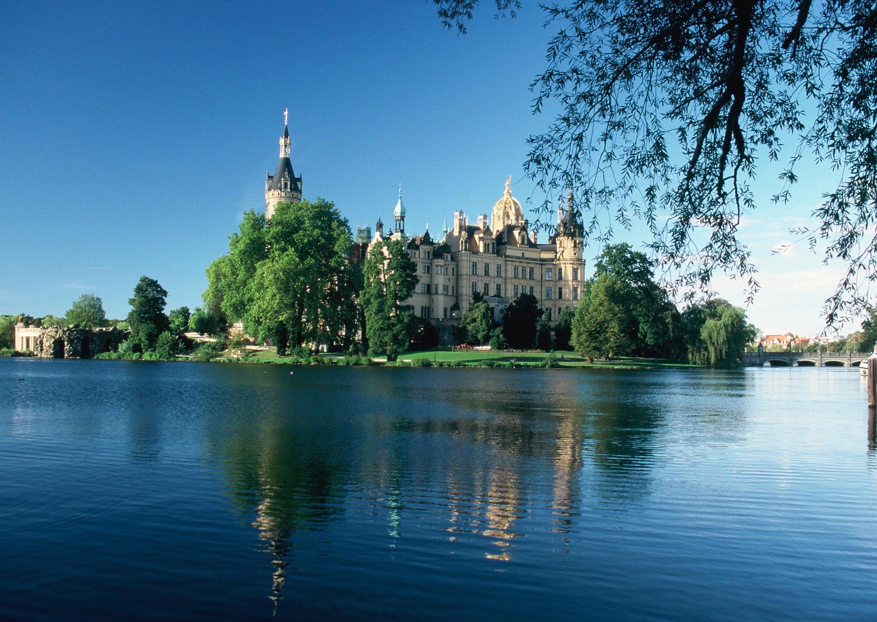 A large, historic building with multiple towers and domes, surrounded by lush greenery and reflecting on a calm body of water.