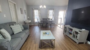 A living room with a grey sofa, a coffee table, a TV stand, and a dining area with a table and chairs. - Cozy home featuring a screened-in porch and access to a refreshing pool. Your oasis awaits!! (Ocean Pines)