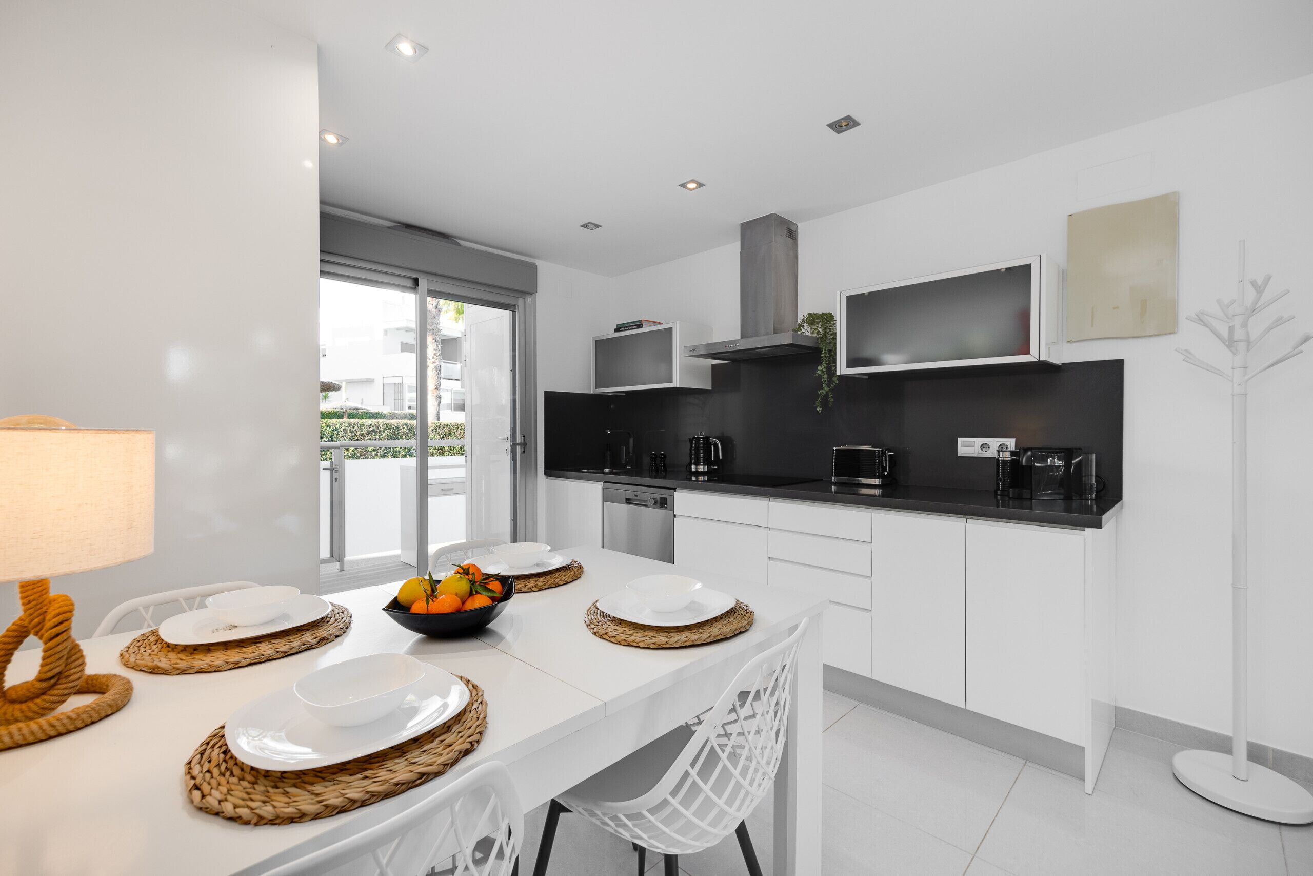 A modern kitchen with white cabinetry, black countertops, and a dining area with white chairs and woven placemats.