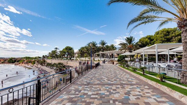 A beachside walkway with a tiled path, palm trees, and outdoor seating areas. - Groundfloor Amay Punta Prima pool/sauna (Torrevieja)