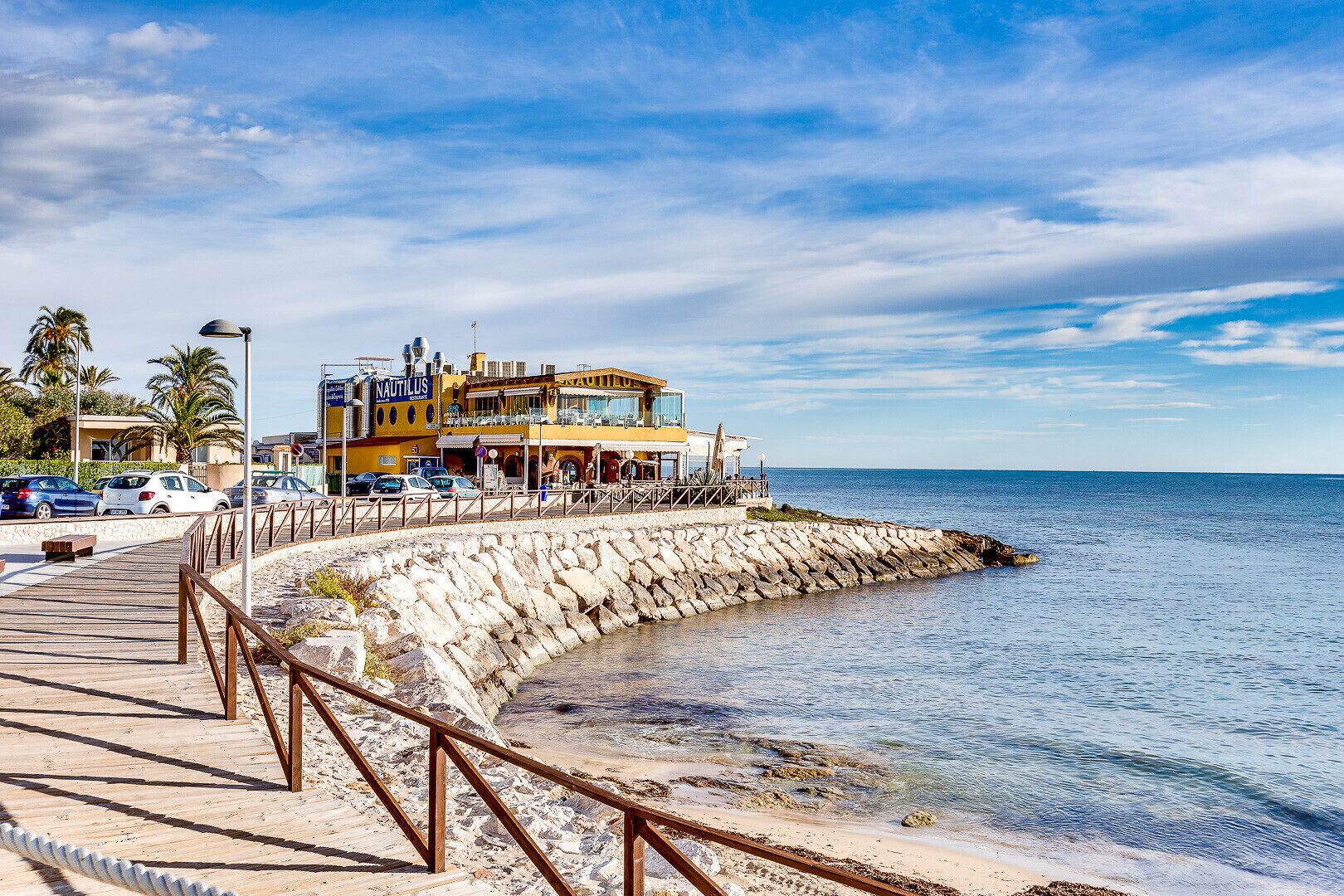 Un hotel frente al mar con vistas, un paseo marítimo de madera y un rompeolas rocoso.