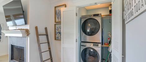 A laundry room with two stacked front-loading washing machines, a wooden ladder, a white door, and a flat-screen TV mounted above a fireplace.