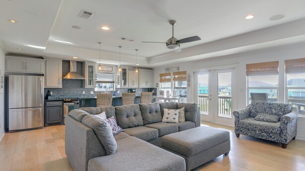 A spacious living area with a gray sectional sofa, a patterned armchair, and a kitchen with white cabinets and a stainless steel refrigerator.