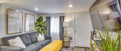 A modern living room with a grey sofa, a yellow ottoman, a TV, and a plant.
