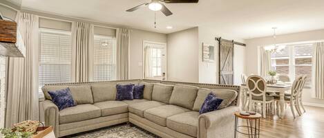 A living room with a sectional sofa, a dining area with a table and chairs, a ceiling fan, and a patterned rug.