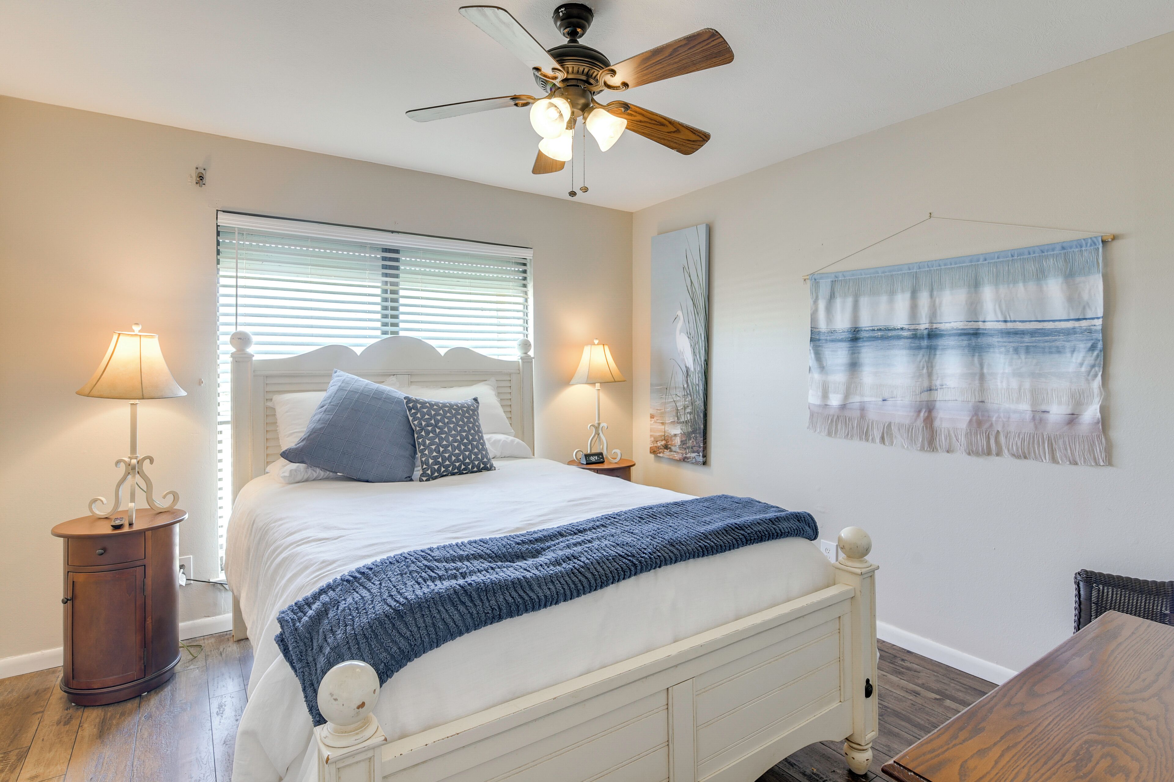 A bedroom with a bed, two bedside lamps, a ceiling fan, a window with blinds, and a wall-mounted tapestry.