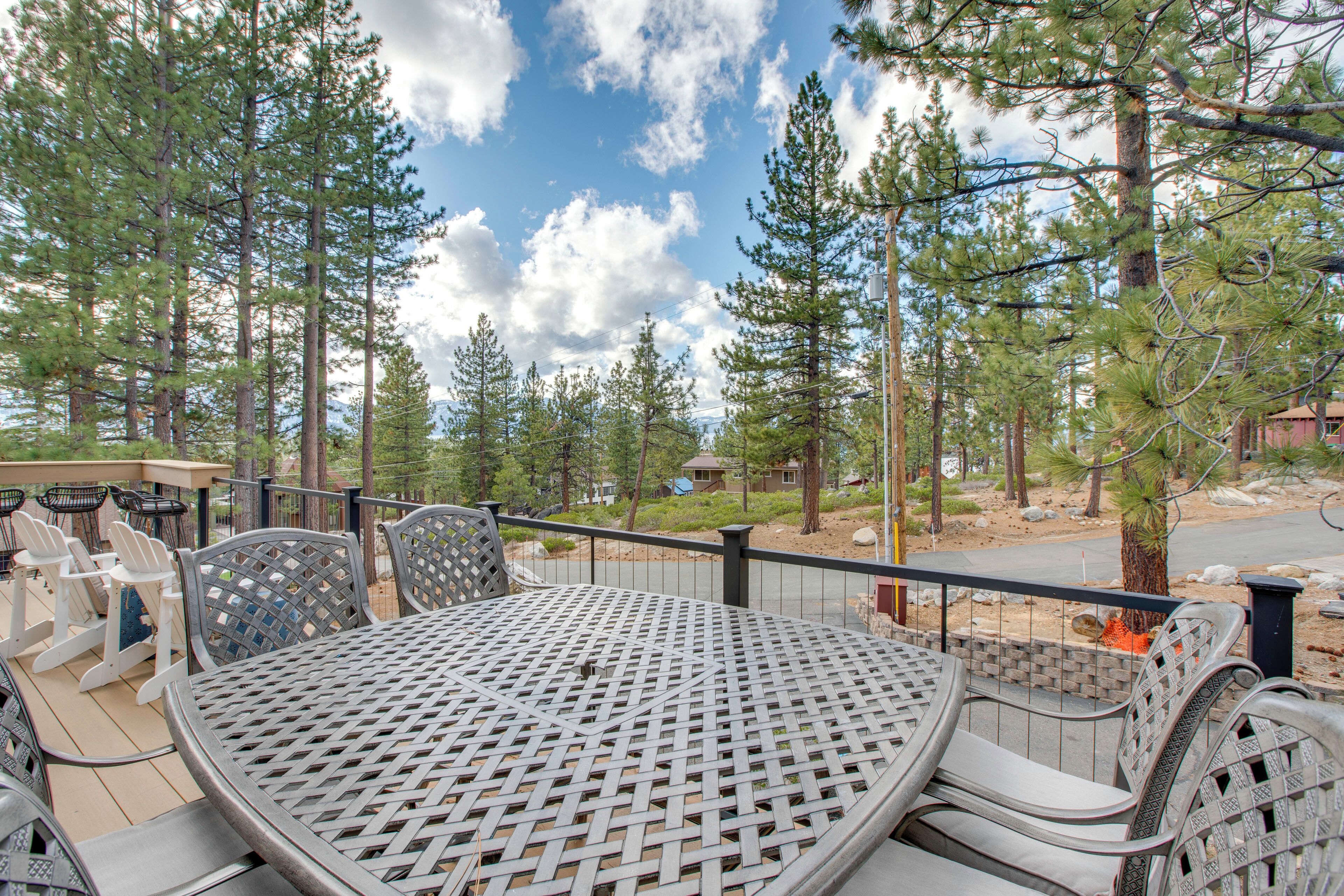 Une terrasse extérieure avec une table et des chaises rondes en métal, entourée de grands pins et sous un ciel dégagé.
