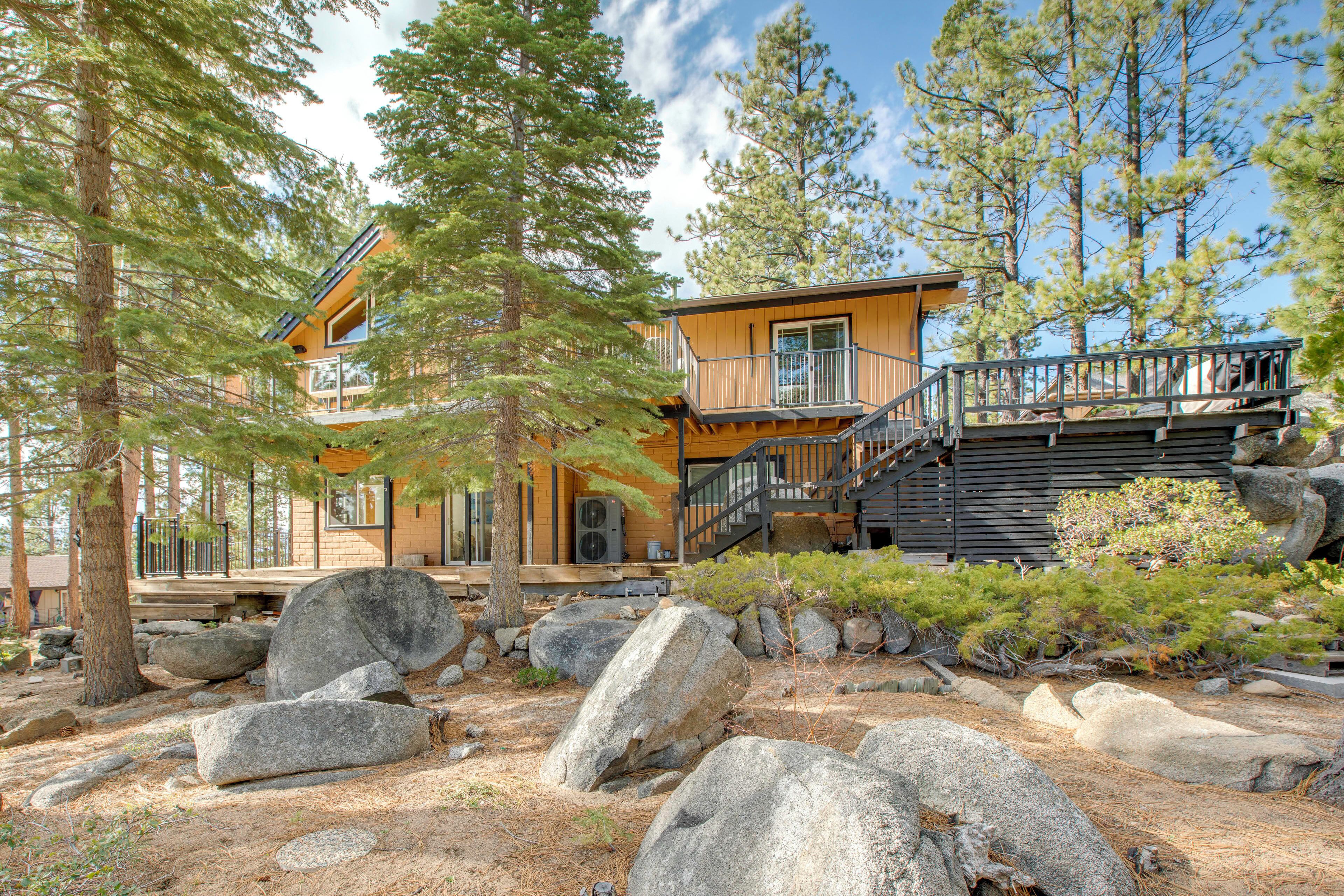 A two-story wooden cabin with a balcony, surrounded by large rocks and pine trees.