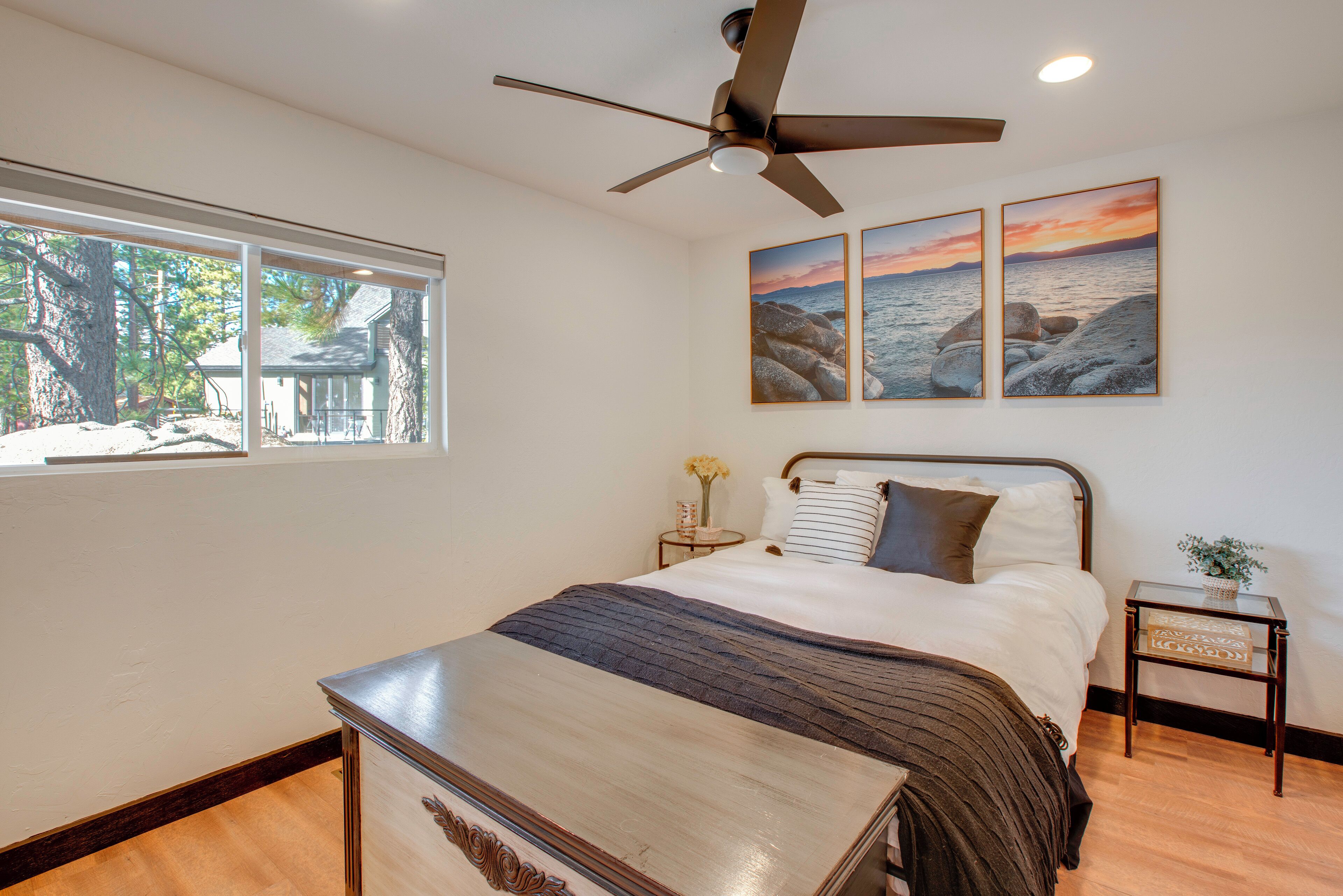 Une chambre à coucher avec un lit, un ventilateur de plafond, une fenêtre donnant sur des arbres et deux tableaux encadrés accrochés au mur.