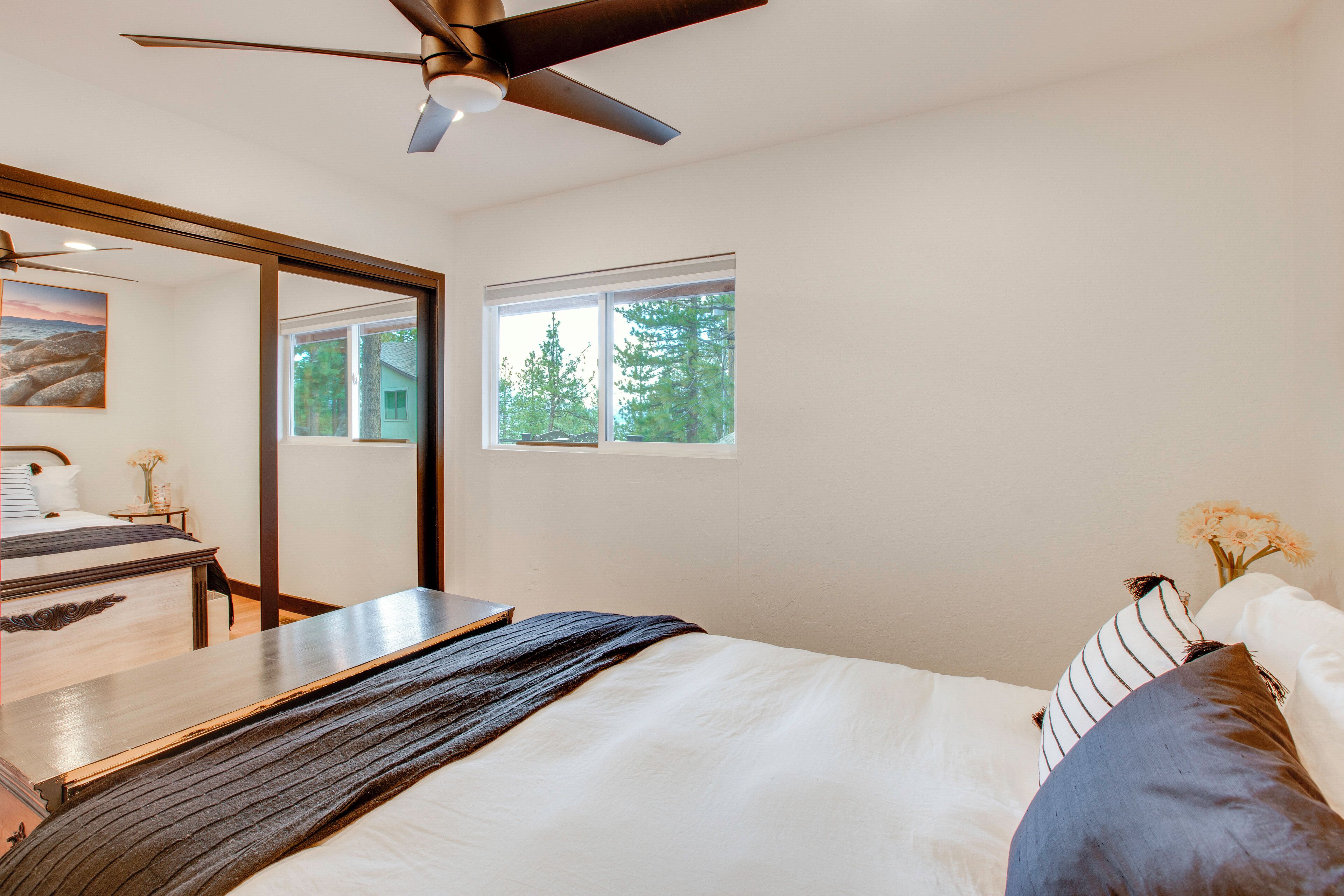 Un dormitorio con cama, escritorio, silla, ventilador de techo y ventana con vistas a árboles.