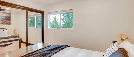 Un dormitorio con cama, escritorio, silla, ventilador de techo y ventana con vistas a árboles.