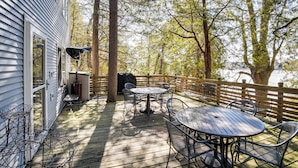 Une terrasse en bois, meublée pour l’extérieur, avec un barbecue et une vue sur les arbres et un bâtiment.