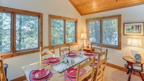 Un coin repas avec une table en bois dressée pour quatre personnes, entourée de chaises en bois, de grandes fenêtres donnant sur les arbres et une lampe posée sur une table d’appoint.