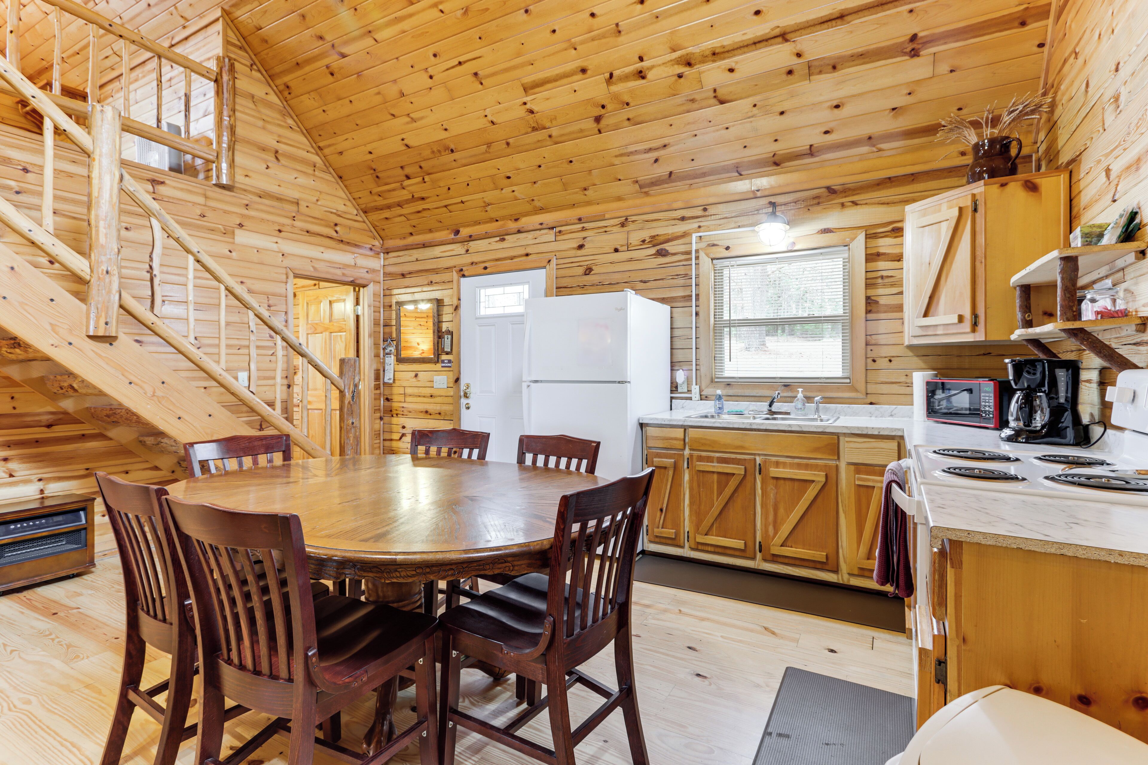 A wooden cabin interior with a dining table, chairs, kitchen area, and a staircase.