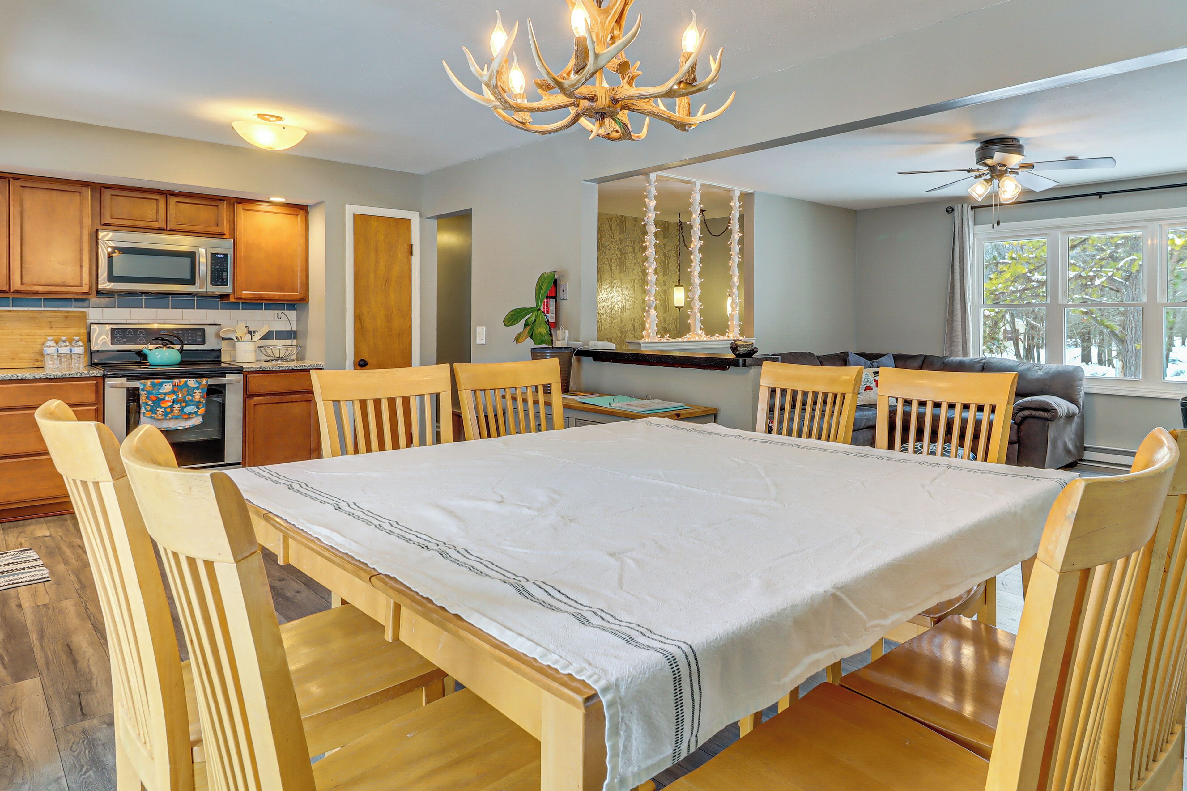 Un espace repas comprenant une table, des chaises, un lustre, un ventilateur de plafond et, au fond, une cuisine.