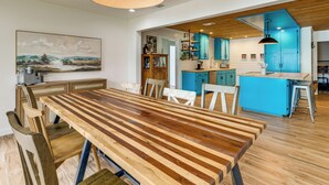 Un espace repas avec une longue table en bois et des chaises, une cuisine avec des meubles bleus et un luminaire suspendu.