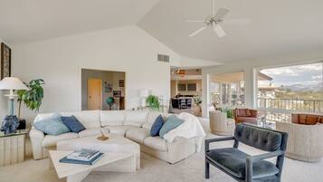 A modern living room with a sectional sofa, a coffee table, and a dark armchair. There is a ceiling fan, a dining area, and a view of the mountains.
