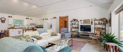 A spacious living area with a sofa, coffee table, and a television. There is a kitchen in the background with cabinets and a window.