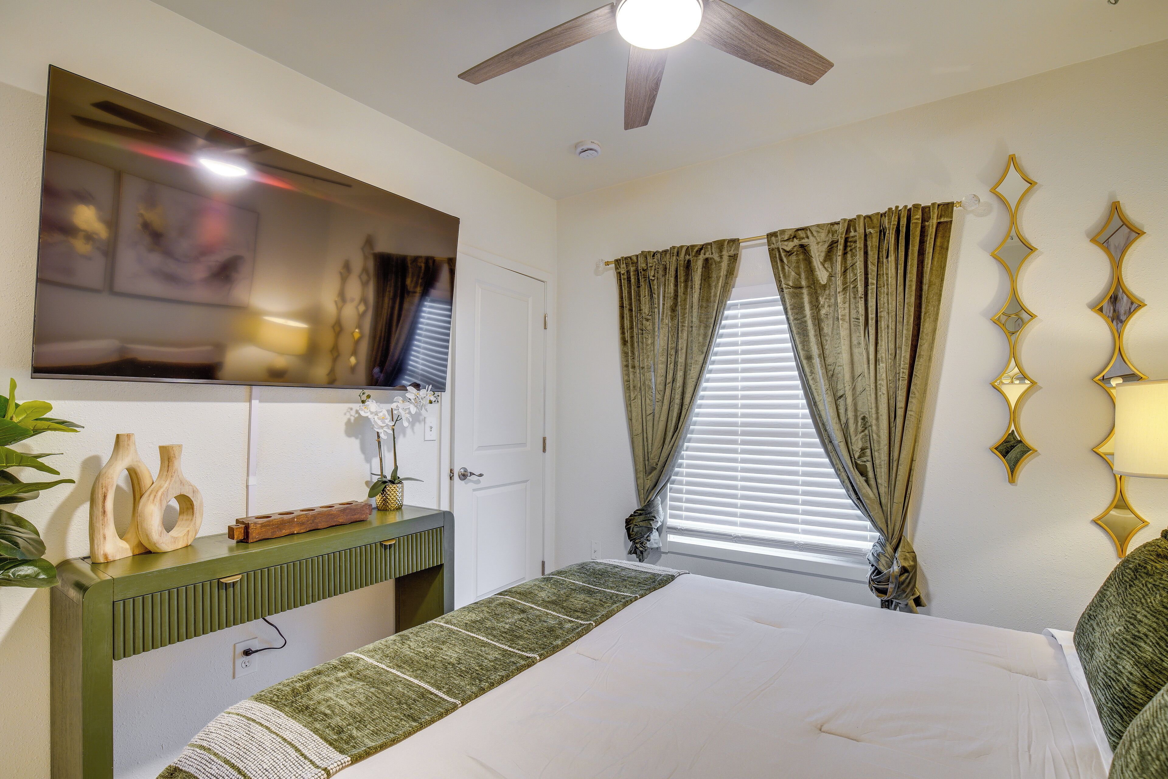 A bedroom with a bed, a ceiling fan, a mounted TV, a bedside table with a plant, and a window with curtains.