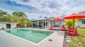 Piscina com área de varanda coberta, cadeiras Adirondack vermelhas e um pequeno edifício com fachada branca.