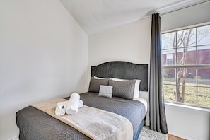 Una cama bien hecha con cabecera, una ventana con cortinas y vista a un edificio de ladrillo.