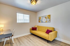 A room with a yellow sofa, a dining table with chairs, a window with blinds, and a wall-mounted light fixture.