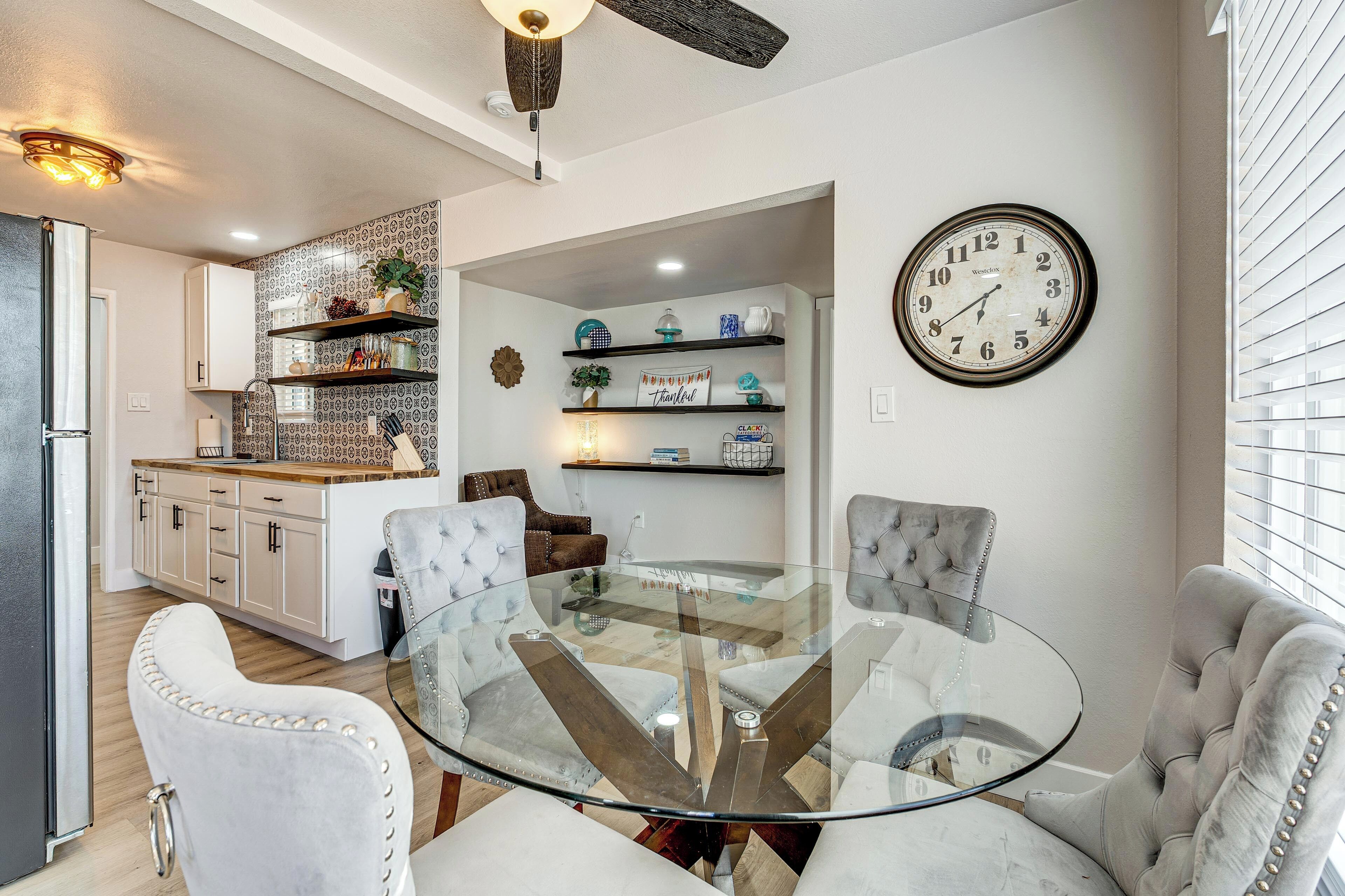 A dining area with a glass table, cushioned chairs, a kitchen in the background, and a wall clock.