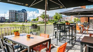 An outdoor patio with tables and chairs, umbrellas, and a view of buildings in the background. - Make Your Trip More Enjoyable! 2 Relaxing Units, Onsite Parking and Dining! (Helmond)