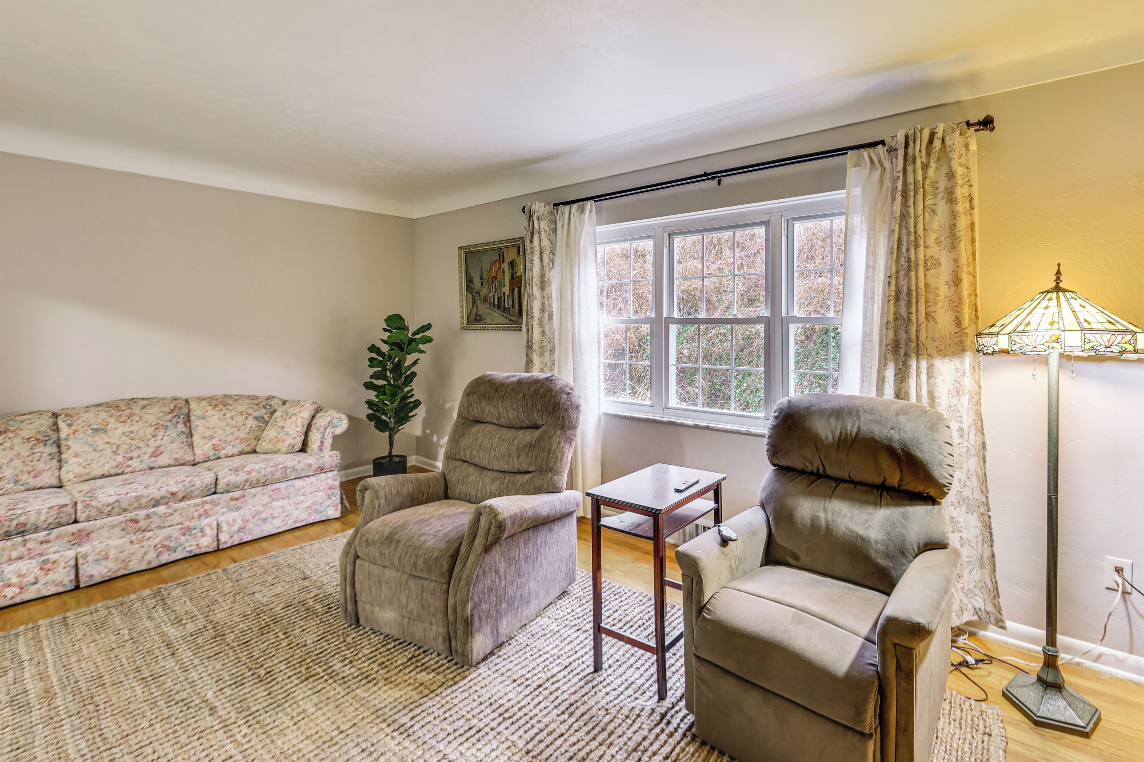 A living room with a sofa, two armchairs, a small table, a floor lamp, a potted plant, and a window with curtains.