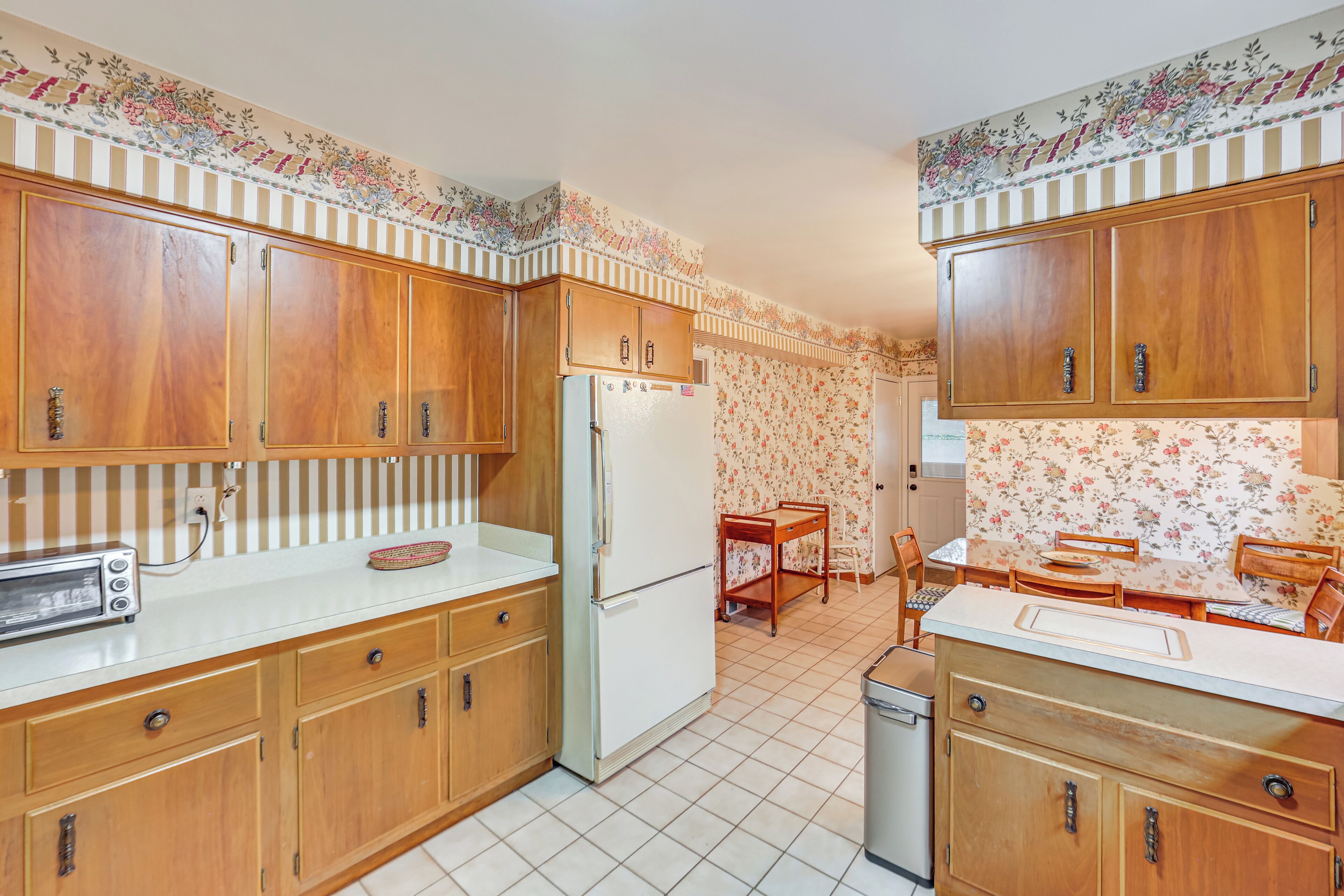 A kitchen with wooden cabinets, a refrigerator, a dining table with chairs, and a tiled floor.